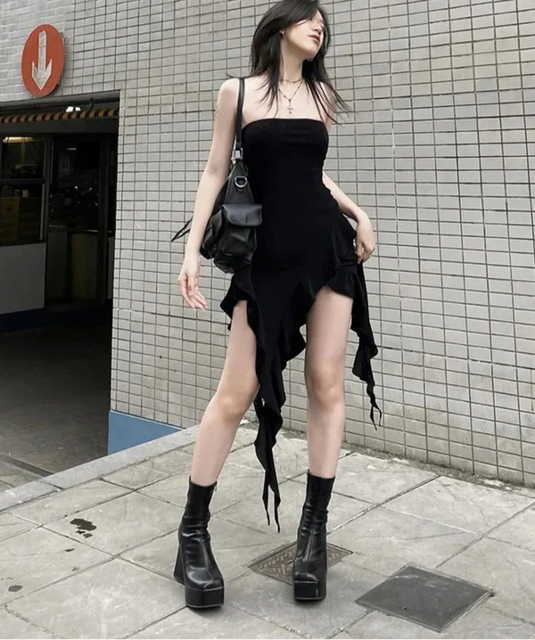 Woman in a black dress and boots standing against a tiled wall.