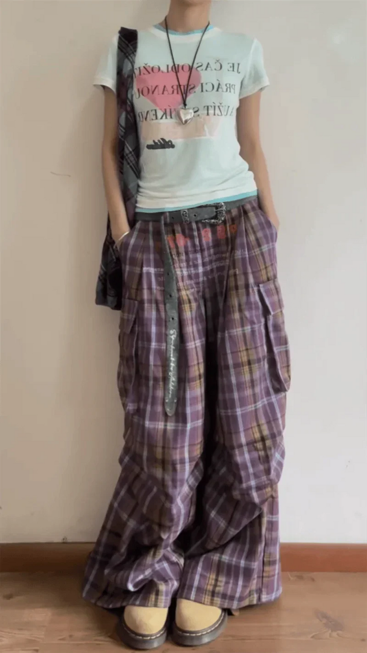 Person wearing a light-colored t-shirt with text and plaid pants against a plain wall.
